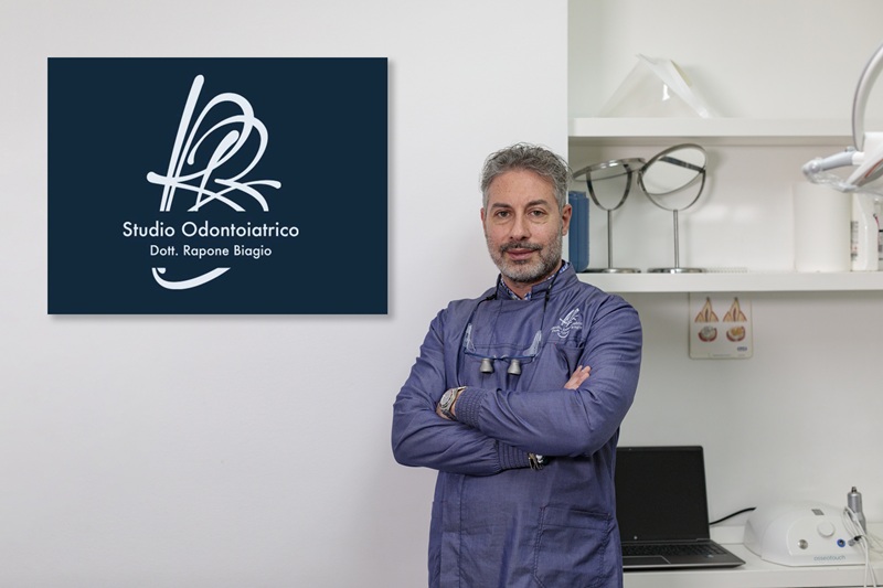 Dentista in casacca blu con occhialini ingranditori accanto all'insegna Studio Odontoiatrico Dott. Rapone Biagio
