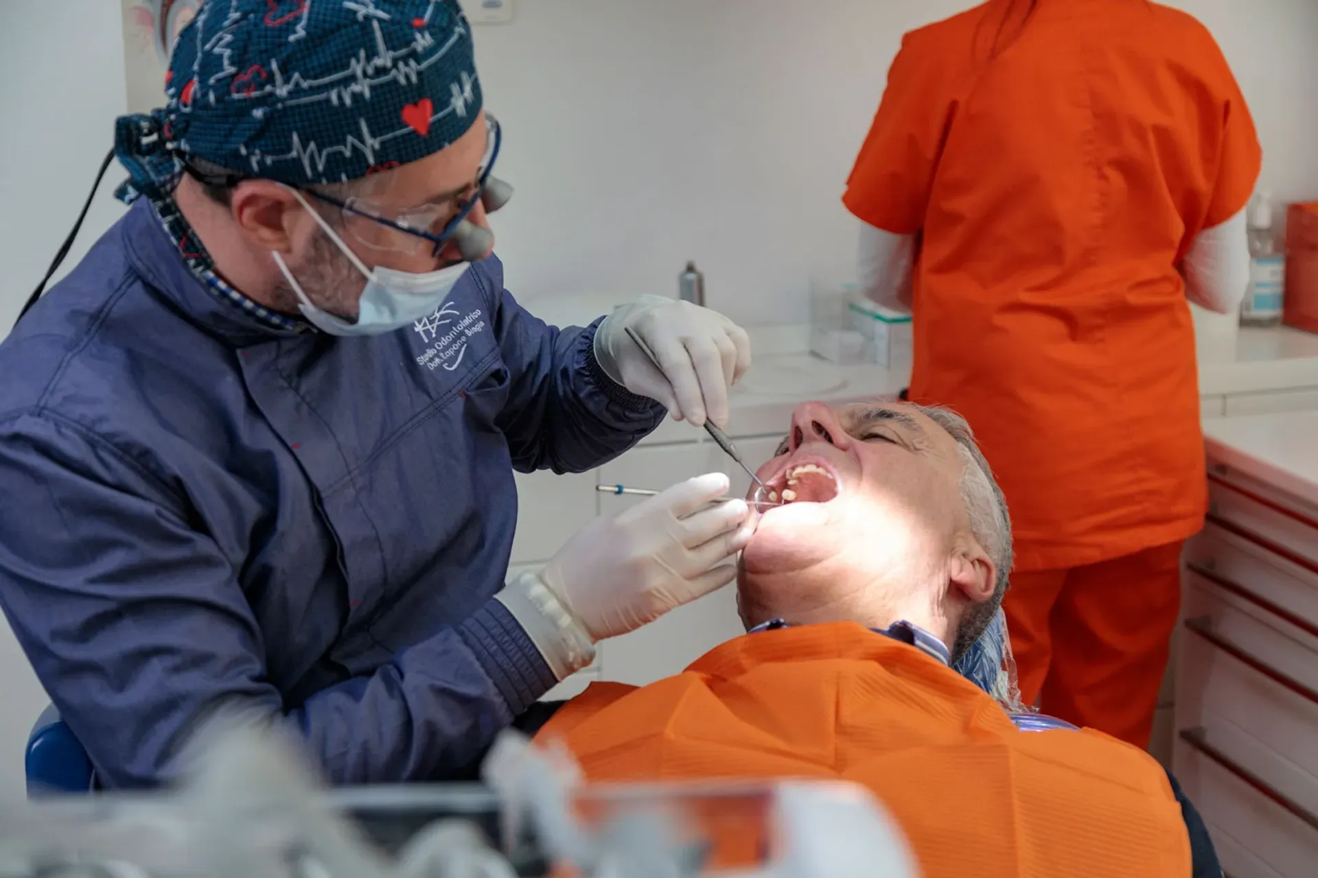 Dentista con lenti ingrandenti e cuffia a cuori visita un paziente con bavaglino arancione, studio Dott. Biagio Rapone